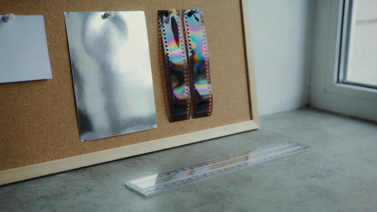 Corkboard with a silver sheet, iridescent film strips, and a clear ruler on a desk